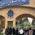 نمای ورودی اصلی ساختمان مدرسه با معماری سنتی خاکی و دروازه‌های فلزی سبز، تابلوی نام مدرسه در بالا