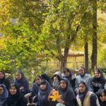 گروهی از دانش‌آموز دختر پوشش‌یافته به حجاب در حیاط سایه‌دار درختان، در حال برگزاری یک فعالیت یا رویداد گروهی