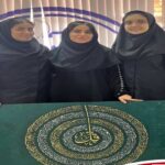 سه دانش‌آموز دختر در حال نمایش پروژه قرآنی با تابلوی سبز رنگ و متن طلایی قرآنی