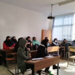 دانش‌آموزان در یک کلاس درس نشسته اند و درس می‌شنوند. میزها و صندلی‌های چوبی، نور طبیعی از پنجره‌های بزرگ، محیط آموزشی داخلی.
