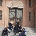 نمای بیرونی سردر و درب اصلی ورودی ساختمان مدرسه با معماری سنتی، دانش‌آموزان نشسته روی پلکان گرانیتی