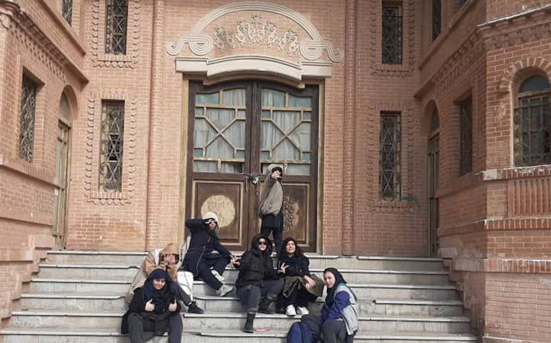 نمای بیرونی سردر و درب اصلی ورودی ساختمان مدرسه با معماری سنتی، دانش‌آموزان نشسته روی پلکان گرانیتی