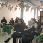 گردهمایی بانوان در سالن درونی مزین شده با پرچم‌های رنگی، میز پذیرایی و فضای اجتماعی