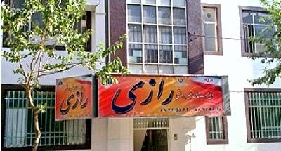 نمای جلوی ساختمان اصلی مدرسه با تابلوی قرمز و درهای سبز، احاطه شده توسط درختان سبز و آسمان ابری