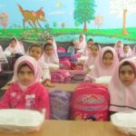 گروهی از دانش‌آموزان دختر در لباس مدرسه و حجاب وردی رنگ در کلاس درسی نشسته‌اند. دیوار پشت آن‌ها با نقاشی رنگی از حیوانات و درختان تزیین شده است و حقائب مدرسی وردی روی میزها قرار دارد.