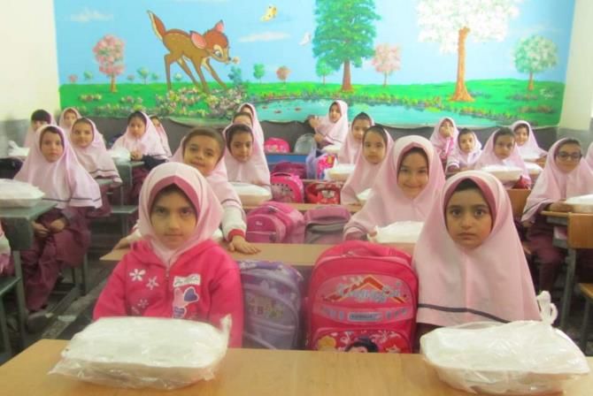 گروهی از دانش‌آموزان دختر در لباس مدرسه و حجاب وردی رنگ در کلاس درسی نشسته‌اند. دیوار پشت آن‌ها با نقاشی رنگی از حیوانات و درختان تزیین شده است و حقائب مدرسی وردی روی میزها قرار دارد.