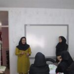 یک معلم زن در لباس زرد ایستاده در کنار سفیدتخته در حال تدریس به دانش‌آموزان