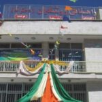 نمای بیرونی ساختمان مدرسه‌ای سفید رنگ با طبقات متعدد، پنجره‌ها، تابلوهای راهنما و دکوراسیون روز ملی
