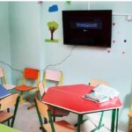 کلاس درس پیش‌دبستان با میزهای نقالی رنگی و صندلی‌های نارنجی، دیوارهای فیروزه‌ای تزئین‌شده با تصاویر و کاردستی‌های کودکانه، و صفحه نمایش تلویزیونی