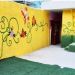 حیاط مدرسه با دیوارهای رنگی زرد و سبز، نقاشی‌های گل و گیاهان تزئینی رنگارنگ، درب‌های آبی و فضایی شاداب برای بازی و تفریح دانش‌آموزان