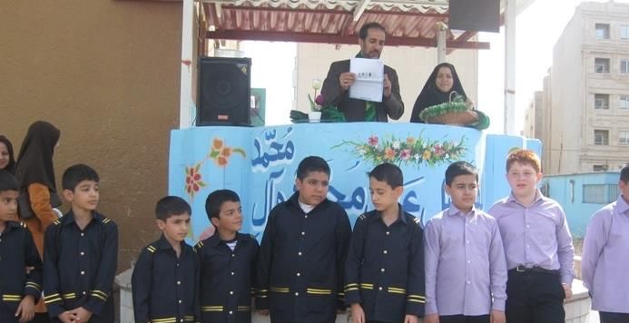 دانش‌آموزان شرکت‌کننده در یک رویداد مدرسه‌ای با بنر آبی و سرپرستان در فضای باز تحت چتری