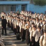 دسته‌ای از دانش‌آموزان در لباس یکسان سفید و قهوه‌ای در حیاط مدرسه در حین یک مراسم یا فعالیت گروهی تحت نظارت اساتید
