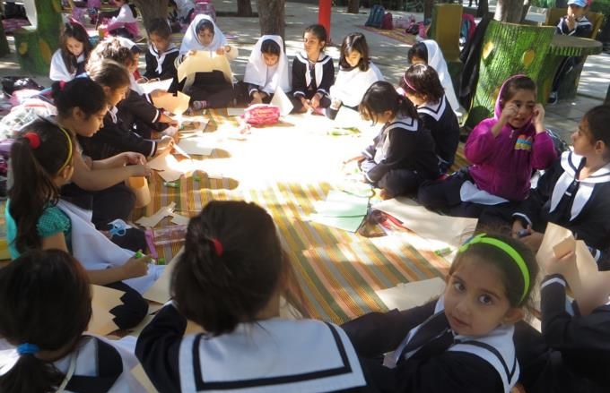 دانش‌آموزان دختری در حیاط اطراف یک پارچه بزرگ نشسته‌اند و در یک فعالیت/پروژه گروهی شرکت می‌کنند.