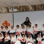 گروهی از دانش‌آموزان دختر با لباس سفید در یک مراسم یا اجتماع مدرسه‌ای، تزیین شده با بالونهای قرمز در یک سالن داخلی