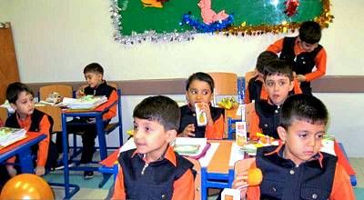 دانش‌آموزان در حال انجام فعالیت گروهی و تیمی در کلاس درس، نشسته دور میز‌های نارنجی با لباس مدرسه