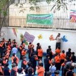 گروهی از دانش‌آموزان در حیاط مدرسه در حال شرکت در یک رویداد جشن با بنرهای رنگی تزیینی در پس‌زمینه