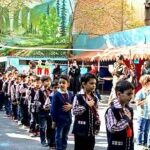 گروهی از دانش‌آموزان مدرسه که در صفوف منظمی در حیاط ایستاده‌اند، حین یک مراسم یا رویداد جمعی، با چتری حفاظتی آبی و درختان سبز در پس‌زمینه