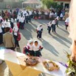 صحنه‌ای از رویداد جمعی در حیاط مدرسه، با صدها شرکت‌کننده، میز‌های خوراکی، و نور طبیعی روز