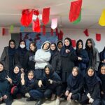 گروهی از دانش‌آموزان دختر در یک فعالیت مراسمی با پرچم‌های رنگی (قرمز، سبز، زرد) که در سقف تزیین شده‌اند