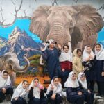 گروهی از دانش‌آموزان در حال عکس‌گیری جمعی جلوی دیوار نقاشی شده‌ای با تصویر فیل، کوه، درختان و آسمان آبی