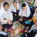 گروهی از دختران دانش‌آموز با حجاب سفید و یونیفرم تیره در فضای باز (حیاط) نشسته‌اند و یک جشن غذایی یا پیکنیک را برگزار می‌کنند