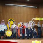 گروهی از دانش‌آموزان در یک اجرای تئاتری مدرسه‌ای روی صحنه‌ی دارای دیوارهای چوبی، با لباس‌های مدرسه و تجهیزات صحنه‌ای اجرای فصلی.