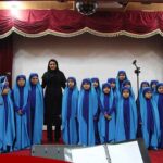 دانش‌آموزانی پوشیده‌ی دیباچه‌ی آبی در حین اجرای موسیقی روی صحنه‌ی تزیین‌شده با پرده‌های قرمز و مایکروفون