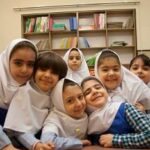 گروهی از دانش‌آموزان دختر با روسری سفید در کلاس درس، کنار یکدیگر نشسته‌اند و برای عکس پوز می‌دهند، قفسه‌ی کتاب‌های درسی در پس‌زمینه قابل‌رویت است