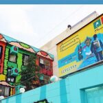 نمای خارجی مدرسه با پیشروی ملون‌شده در رنگ‌های سرخ، سبز، آبی و زرد، با رسوم‌آنی و لافتۀ آموزشی روی دیوار