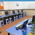کلاس سایت کامپیوتری مجهز به میز‌های آبی و رایانه‌های رومیزی برای آموزش تکنولوژی و مهارت‌های دیجیتال