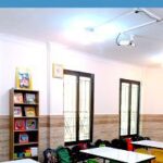 کلاس درسی مجهز با میز‌های رنگی، قفسه‌های کتاب و وسایل آموزشی تحت نور طبیعی
