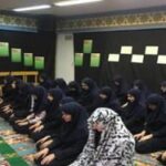 دانش‌آموزان در حال نماز در نمازخانه مدرسه با تزیینات سنتی و فرش رنگین