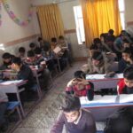 گروهی از دانش‌آموزان در کلاس درس نشسته‌اند در پشت میزهای فردی و در حال انجام فعالیت درسی، دیوار پشت سفید با تزیینات رنگی، پنجره‌های بزرگ با پرده‌های زرد
