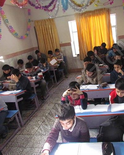 گروهی از دانش‌آموزان در کلاس درس نشسته‌اند در پشت میزهای فردی و در حال انجام فعالیت درسی، دیوار پشت سفید با تزیینات رنگی، پنجره‌های بزرگ با پرده‌های زرد