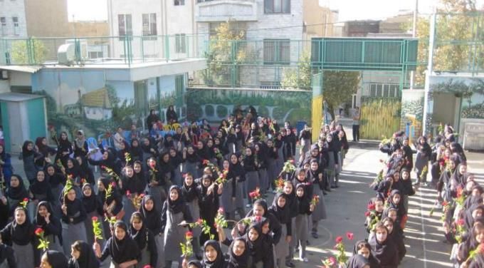 دانش‌آموزانی در پوشش یکپارچه در حیاط مدرسه در حال شرکت در مراسمی دسته‌جمعی با گل و گیاه