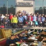 گردهمایی جمعی دانش‌آموزان و اعضای مدرسه در فضای باز جلوی ساختمان؛ گروهی از افراد ایستاده در پشت‌زمین در کنار لوگوی مؤسسه و دیگران نشسته در جلو حول میزی با مواد و تجهیزات گردهمایی