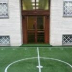 نمای خارجی ساختمان مدرسه با درب ورودی قرمز و پنجره‌های محافظ‌شده، و کورت فوتبال با خطوط سفید در پیش‌زمین