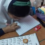 دانش‌آموزی در حال نوشتن و کار روی کاغذ با لوازم مدرسی (ربات، کاغذ، قلم) روی میز