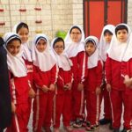 گروهی از دانش‌آموزان دختری با لباس مدرسه قرمز و سفید ایستاده در حیاط مدرسه برای عکس یادیری