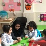 معلمی با حجاب در حال هدایت یک فعالیت آموزشی گروهی برای چند دانش‌آموز دختر در کلاس درس، در حال کار روی میز سبز رنگ با مواد یادگیری