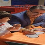 یک دانش‌آموز و معلم در حال کار بر روی پروژه رباتیکی روی میز نارنجی، با ابزارها و تجهیزات فنی