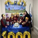 گروهی از دانش‌آموزان و یک معلم در حال جشن با کیک و تزیینات طلایی و آبی، با بالن‌های طلایی حاکی از یک نشان مهم