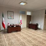 دفتر اداری مدرسه با میز چوبی، پرچم ایران و پرچم مدرسه، تابلوی دیواری، صندلی و تجهیزات اداری
