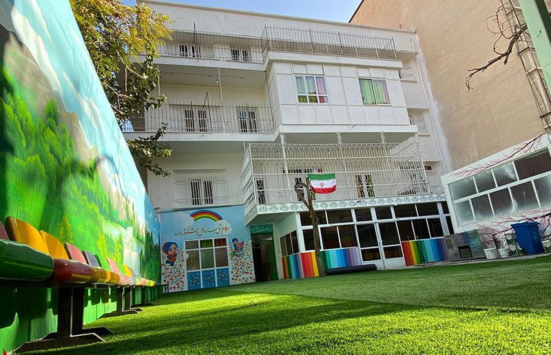 نمای بیرونی ساختمان مدرسه با حیاط سبز، ساختمان سه‌طبقه سفید رنگ و درخت‌های کناری