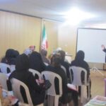 جلسه‌ی رسمی با شرکت‌کنندگان نشسته در صندلی‌های پلاستیکی و سخنرانی مردی ایستاده، پرچم ایران در پس‌زمینه