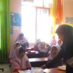 معلم در حال درس دادن به دانش‌آموزان در کلاس درسی روشن و رنگین با صندلی، میز، و تزیینات دیواری