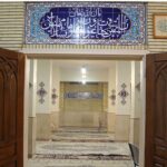 نمازخانه مدرسه با تابلوی فیروزه‌ای رنگ، درب‌های چوبی منقوش، فرش نماز و تزیینات مذهبی