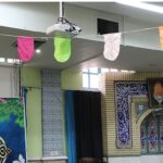 نمازخانه‌ی مدرسه با دیوارهای آراسته به طرح‌های اسلامی و میز‌اب مزین، پرده‌های آبی و نورپردازی سقفی
