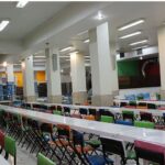 سالن غذاخوری و ناهارخوری با میزهای بلند سفید و صندلی‌های رنگارنگ سبز، قرمز، نارنجی و مشکی، نورپردازی فلورسنت داخلی