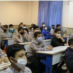 دانش‌آموزان در کلاس درس نشسته‌اند بر روی مقاعد آبی در حال شرکت در درس